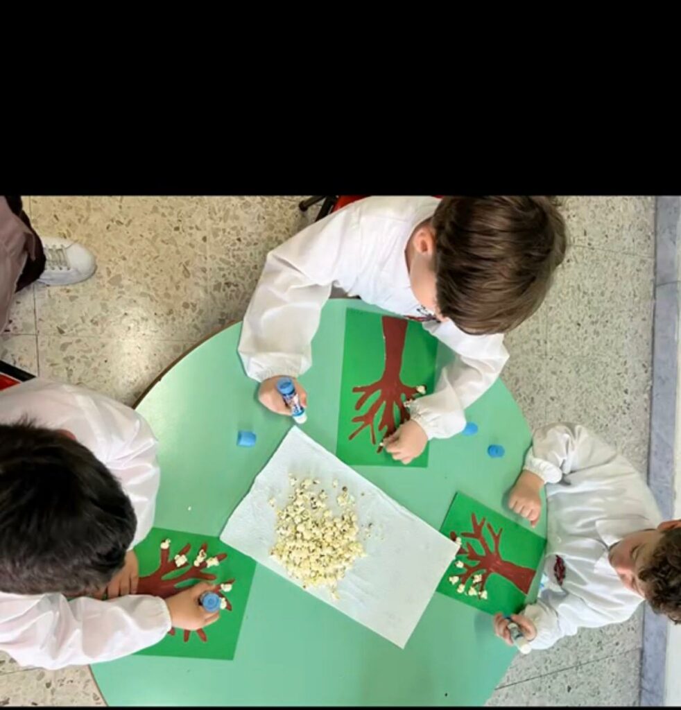 Vista dall'alto di un tavolo rotondo con tre bambini in grembiule che svolgono un'attività creativa, incollando popcorn su disegni di alberi realizzati su cartoncino verde.
