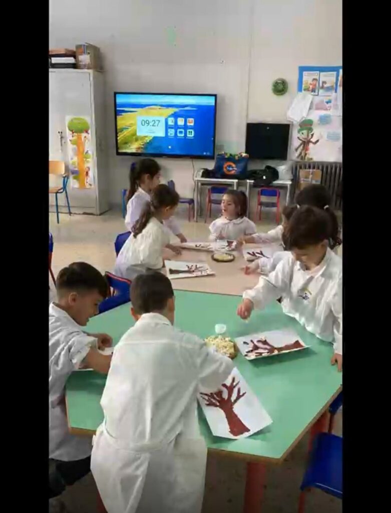 Gruppo di alunni in aula scolastica seduti attorno a tavoli mentre partecipano a un'attività creativa, decorando disegni di alberi con materiali naturali
