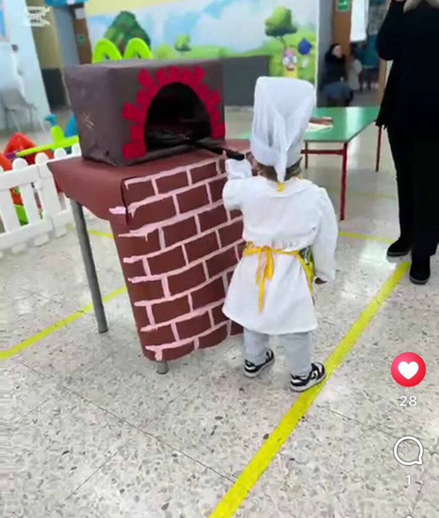 alunno dell'infanzia con cappello e grembiule da cuoco che di spalle inforna una pizza nel forno a legna di cartone 